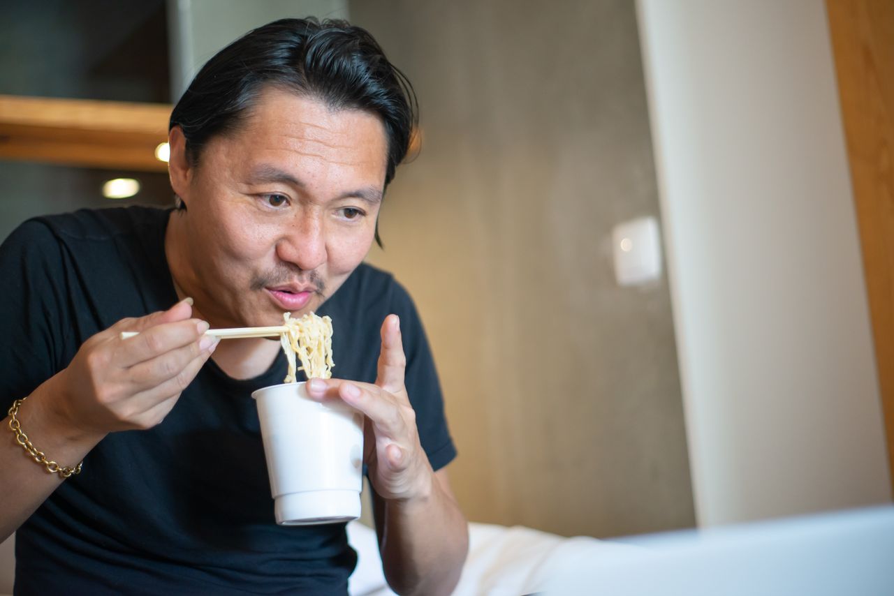 services-04 Asian man sitting indoors enjoying instant noodles with chopsticks in a relaxed setting.